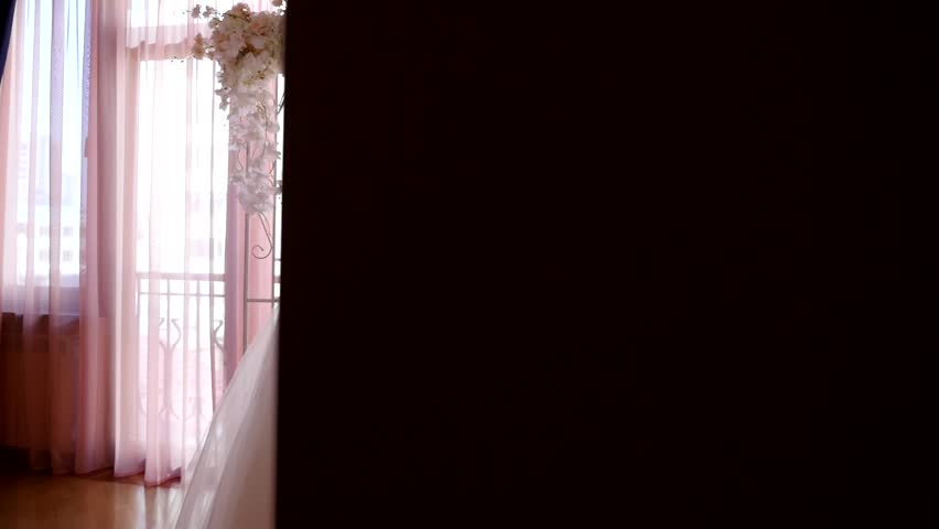 Elegant white wedding dress on mannequin in bright room with floral arch. Bride's hands hold delicate white pouch accessorizing. Ideal for wedding, beauty, fashion, and celebration footage.