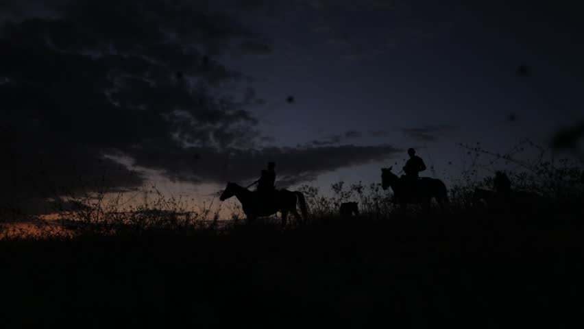 Silhouetted group of people riding horses across a hill against a vibrant sunset sky. Distant mountains and city lights add depth. Evokes freedom, adventure, and natural beauty. Perfect for travel and