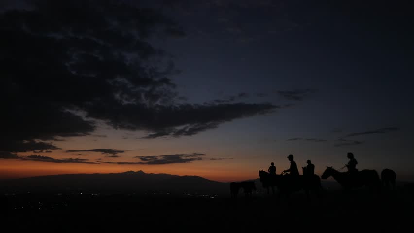 Silhouetted group of people riding horses across a hill against a vibrant sunset sky. Distant mountains and city lights add depth. Evokes freedom, adventure, and natural beauty. Perfect for travel and