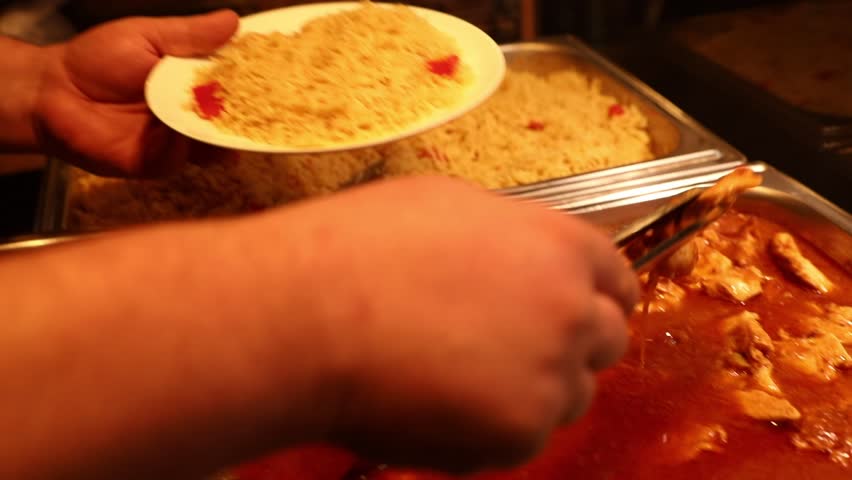 Chef serving rich, red chicken curry from a catering pan. Tongs and ladle scoop out savory pieces and sauce, with rice in background. Ideal for food content.