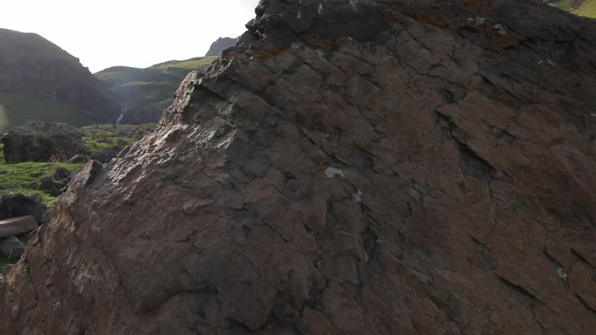 A majestic waterfall cascades down towering mountains, feeding a rushing stream through a lush green valley. Wildflowers adorn rocky slopes, crafting a tranquil natural landscape.