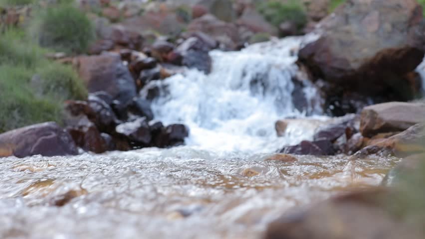A majestic waterfall cascades down towering mountains, feeding a rushing stream through a lush green valley. Wildflowers adorn rocky slopes, crafting a tranquil natural landscape.