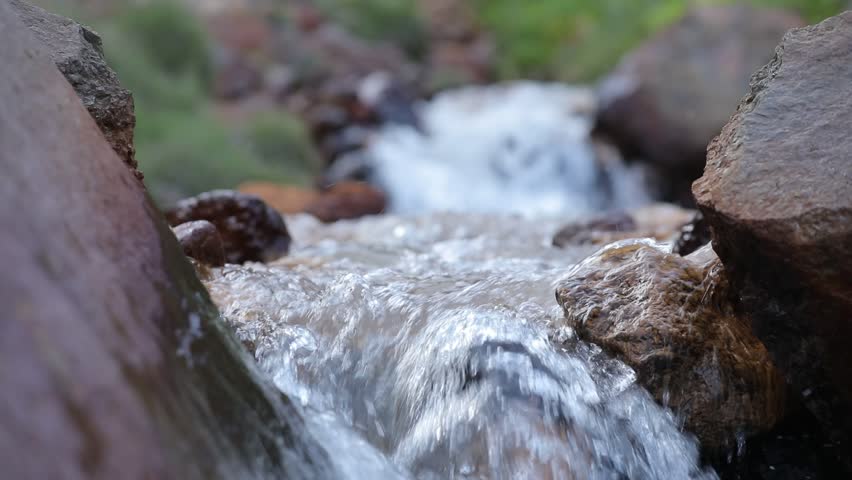 A majestic waterfall cascades down towering mountains, feeding a rushing stream through a lush green valley. Wildflowers adorn rocky slopes, crafting a tranquil natural landscape.