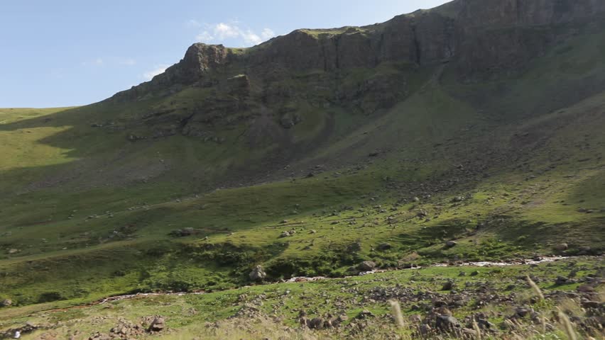 A majestic waterfall cascades down towering mountains, feeding a rushing stream through a lush green valley. Wildflowers adorn rocky slopes, crafting a tranquil natural landscape.