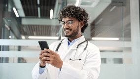 Young smiling doctor in white coat using mobile phone while standing in modern hospital office. Confident handsome medical worker physician in glasses chats online with patient or works in application - Powered by Shutterstock - Get 15% off with code: PIKWIZARD15