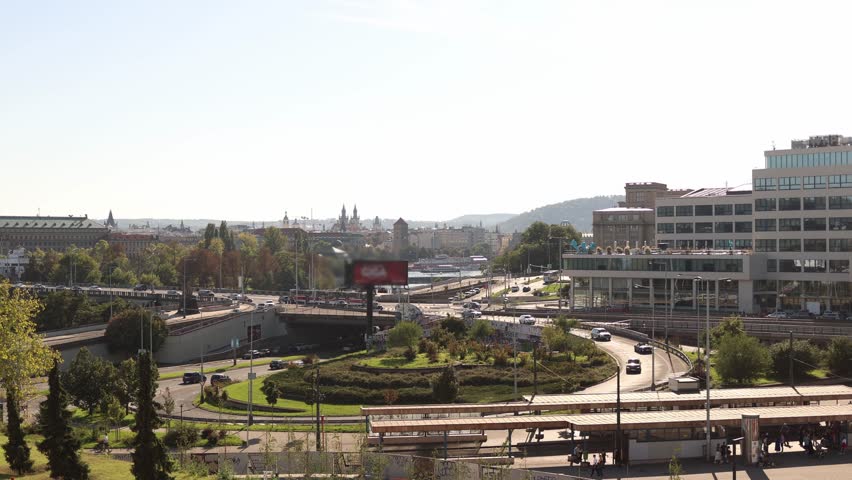 Busy urban roundabout with cars and trams moving on modern roads surrounded by greenery with panoramic view of distant skyline and tall towers in the bright afternoon sunlight