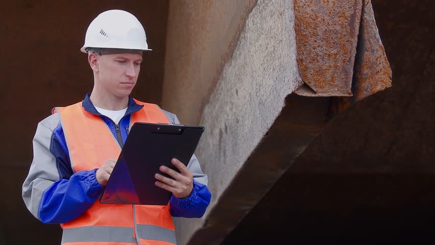 Male dockworker inspecting of hull of ship with clipboard for restoration of iron bottom of barge at shipyard port..