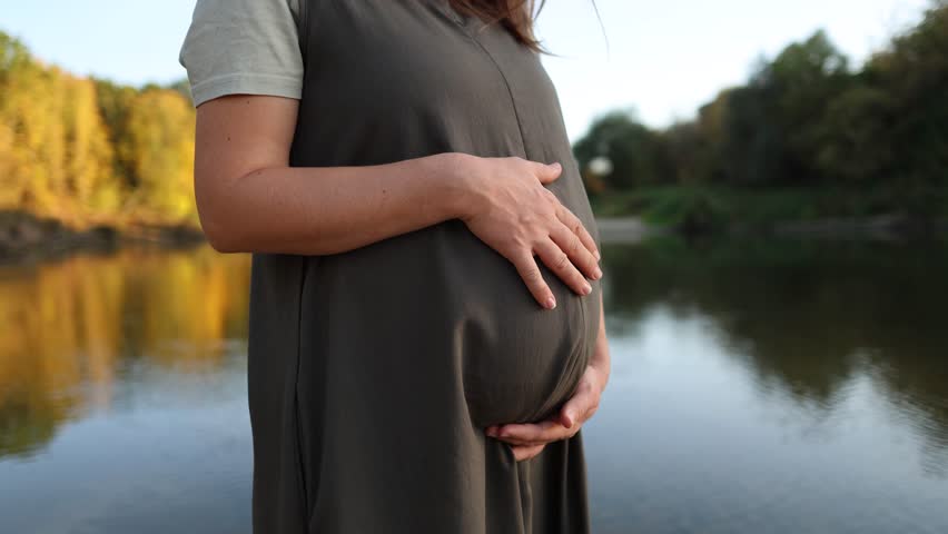 A pregnant Caucasian woman in a green dress walks in a park at sunset. The young mother gently strokes her belly with her hands. The woman is expecting a child.