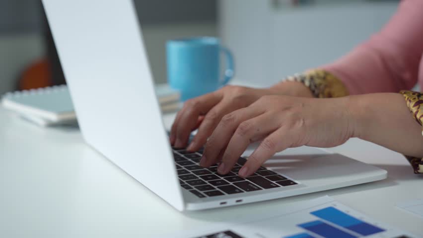Professional businesswoman typing confidentially on laptop keyboard, working intently at office desk with scattered documents, coffee mug, and notepad in close up view