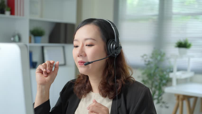 Friendly asian operator woman working in office, she is wearing headset and speaking through microphone, she is smiling and looking at computer screen