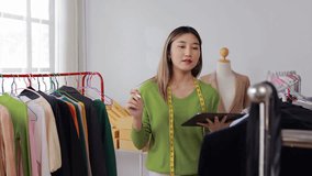 Female fashion designer wearing green sweater working on digital tablet while checking clothing designs and hanging garments in personal studio workshop with natural light - Powered by Shutterstock - Get 15% off with code: PIKWIZARD15