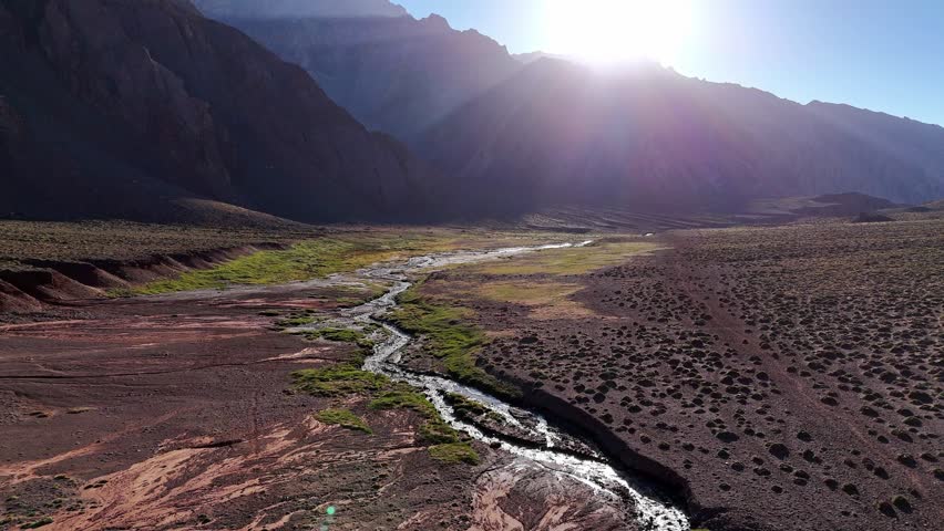 Aerial view of a winding river cutting through the Andes Mountains. The surrounding valley is dry, with green vegetation growing along the water
