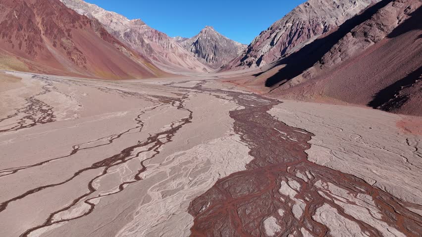 A wide, dry riverbed stretches through the Andes Mountains. The landscape is defined by red and white sediment deposits, with mountains rising in the distance.