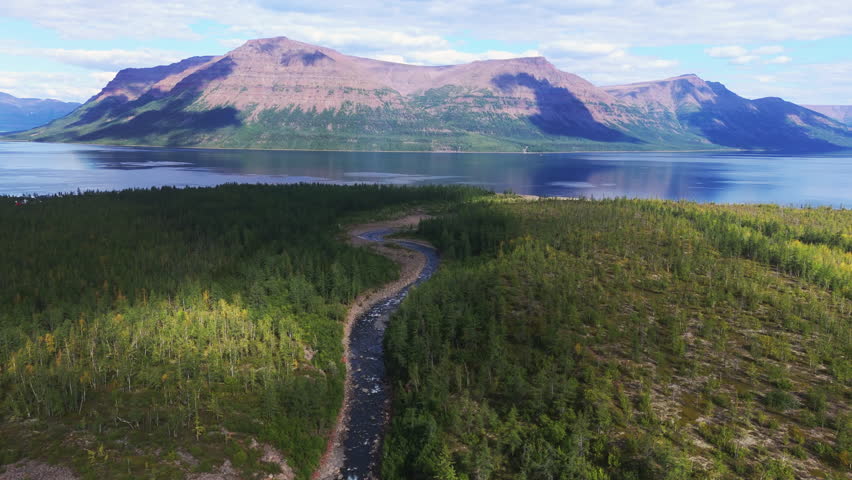 A winding forest river flows toward Lake Lama on the remote Putorana Plateau in Russia. Calm water, mountain reflection and green taiga create an inviting landscape for nature tourism and travel