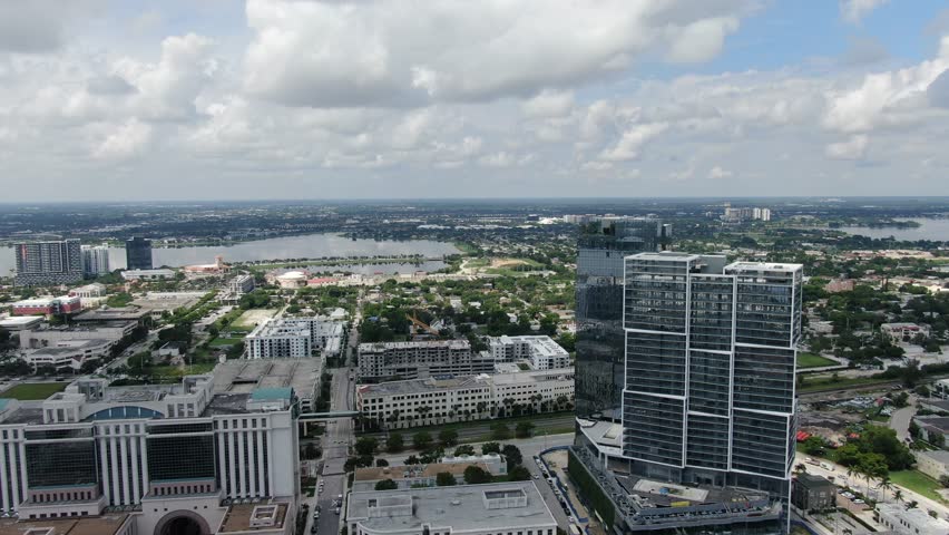 Camera looks west from downtown glass high-rises toward inland lakes and stretches of low-rise neighborhoods in West Palm Beach. Clear daylight and parallax across the urban-to-suburban transition.