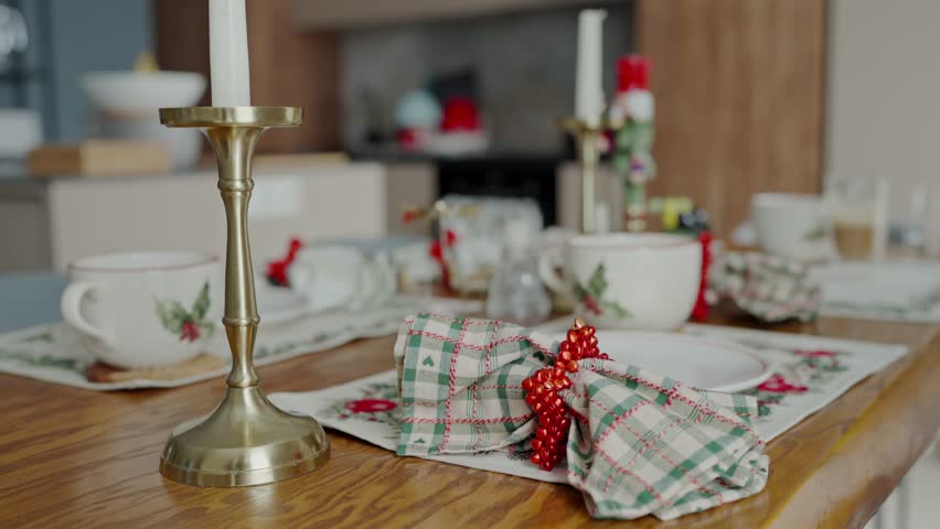 Close-up of a festive Christmas table setting featuring holiday-themed tablecloth, a brass candlestick, and decorative cups. Elegant Christmas Table Setting with Classic Decor and Candle