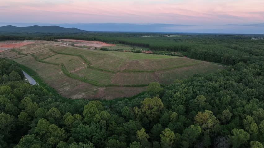 Aerial view of serene landscape featuring rolling hills and expansive fields. Winding road bordered by lush greenery, leading into a tranquil environment. Sunset time in America. Wide shot.