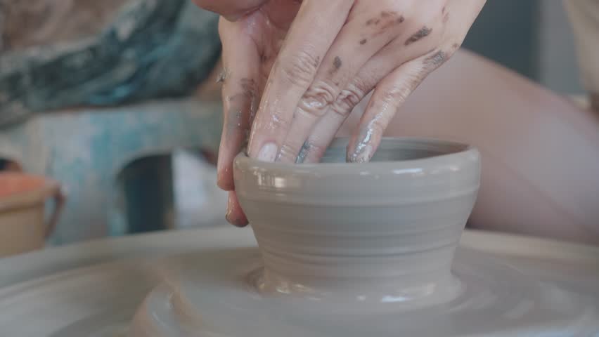 A close-up view of hands skillfully shaping a clay pot on a pottery wheel. The hands display traces of clay, showcasing the art of pottery making and the creative process involved.