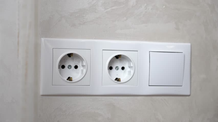 Precise close-up capturing hand inserting white phone charger into dual european wall socket against textured gray wall, focusing on electrical connection process