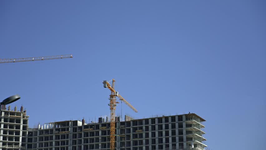 Yellow tower crane moving materials on a large construction site with a new concrete high-rise building being erected against a clear blue sky, showing urban development and housing growth