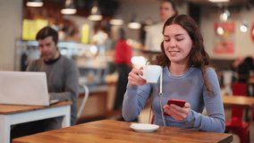 Young woman is sitting at a table in a cafe, drinking hot strong coffee from natural beans and holding a mobile phone in hands. Coffee shop visitor checking social networks on her phone - Powered by Shutterstock - Get 15% off with code: PIKWIZARD15