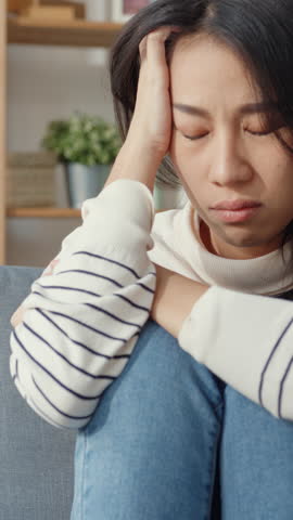 Vertical Screen : Thoughtful Asia lady sit embracing knees at sofa in living room at house look outside with feel lonely, Sad depressed teenager spend time alone stay at home, Social distance concept.