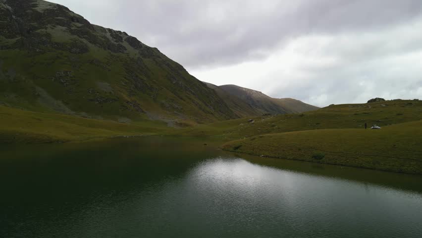 Alpine lake reflecting a grey sky amid grassy hills and a mountain backdrop.