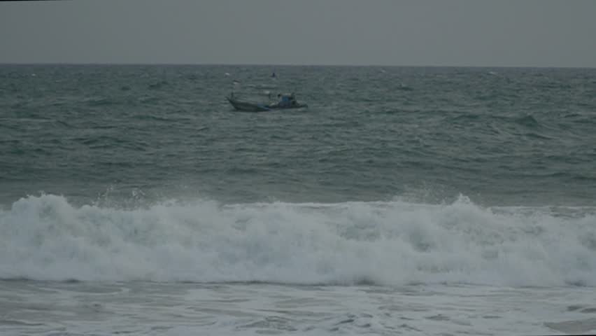 Large, foamy waves crash on the coast under a cloudy sky, with small boats on the horizon, capturing a dramatic and powerful scene of the vast sea. Fishing boats are seen in the middle of the sea 