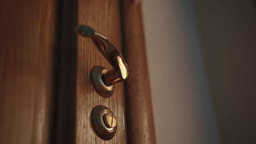 Low-angle view of hand of unrecognizable man gripping golden door handle, signaling imminent movement within softly illuminated hotel room corridor, conveying subtle moment of quiet transition.