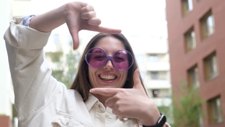 A sweet, cheerful, and charming girl poses for the camera on a city street in the summer. The woman holds up a hand-made frame, like a photograph. A photo frame made by a young hipster girl.
