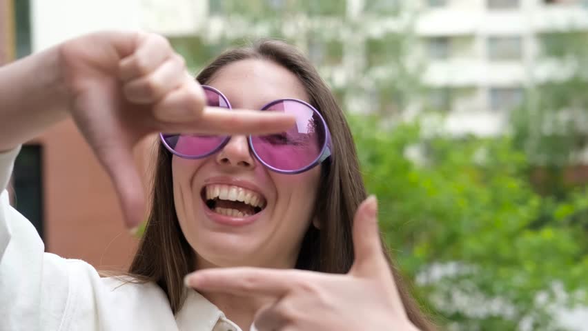 A sweet, cheerful, and charming girl poses for the camera on a city street in the summer. The woman holds up a hand-made frame, like a photograph. A photo frame made by a young hipster girl.
