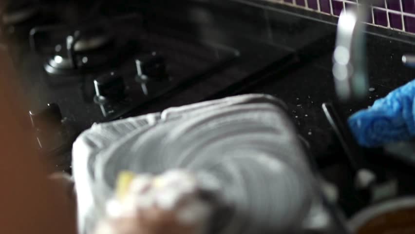 Hand vigorously cleans soiled frying pan in dark kitchen sink, soapy suds covering metal surface. Cleaning tools and blue cloth nearby.