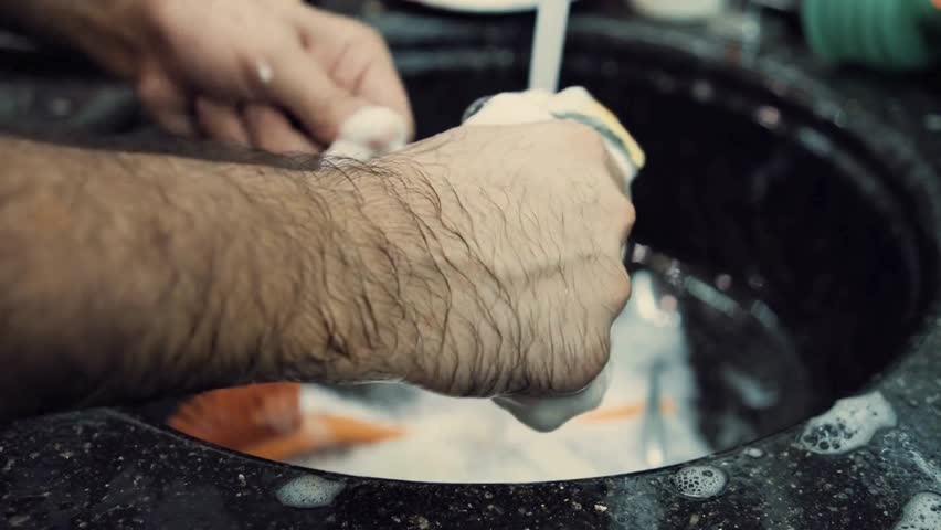 Man washes dishes with sponge in black granite sink, under running water. Lots of suds and bubbles. Orange brush present in sink basin.