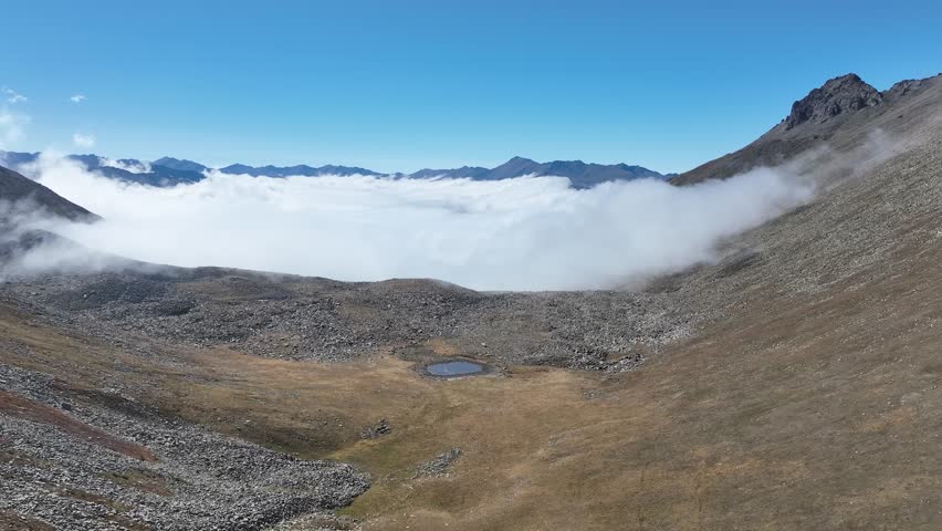 Aerial view of a rocky mountain valley with a small reflective pond, surrounded by ridges and clouds under a clear sky.
