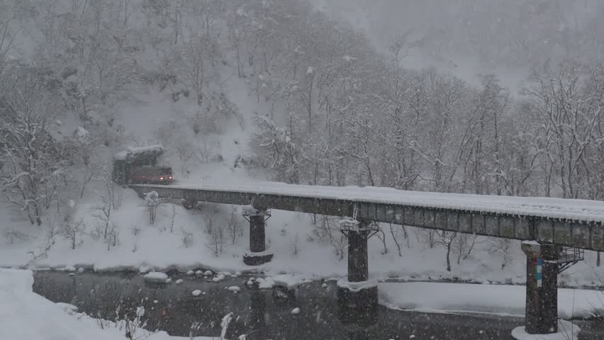 On a cold snowy winter day, a local train of JR Ōito Railway Line comes out of a tunnel and crosses a bridge over Hime-Kawa River in the heavy snowfall, in Otari, Nagano, Japan

Scenic Journey on the Ōito Line
This vibrant image captures a diesel railcar crossing a sturdy iron bridge amidst a heavy winter snowfall in a mountainous region of Japan. The landscape is blanketed in pristine white, the surrounding trees heavily laden with accumulated snow, creating a quiet yet dramatic environment. The dark waters of the Hime River flow beneath the structure, providing a sharp contrast to the bright snow cover. A lone, orange-striped train car (likely a KiHa 120 model) emerges from a nearby tunnel portal, making its solitary passage across the elevated track. The persistent falling snow blurs the background, lending an ethereal quality to this picturesque scene.