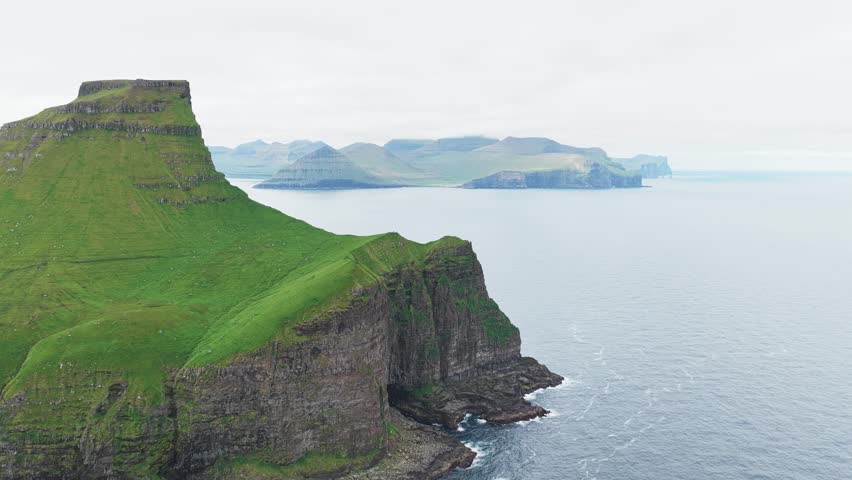 Aerial view of the Kallur Lighthouse standing on a dramatic cliff edge, contrasting with the calm ocean waters and distant islands, Kallur Lighthouse, Norðoya, Faroe Islands.