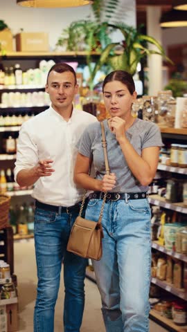 Carefree cheerful young couple engaging in shopping, walking down aisle of organic foods store, exploring with interest shelves with products