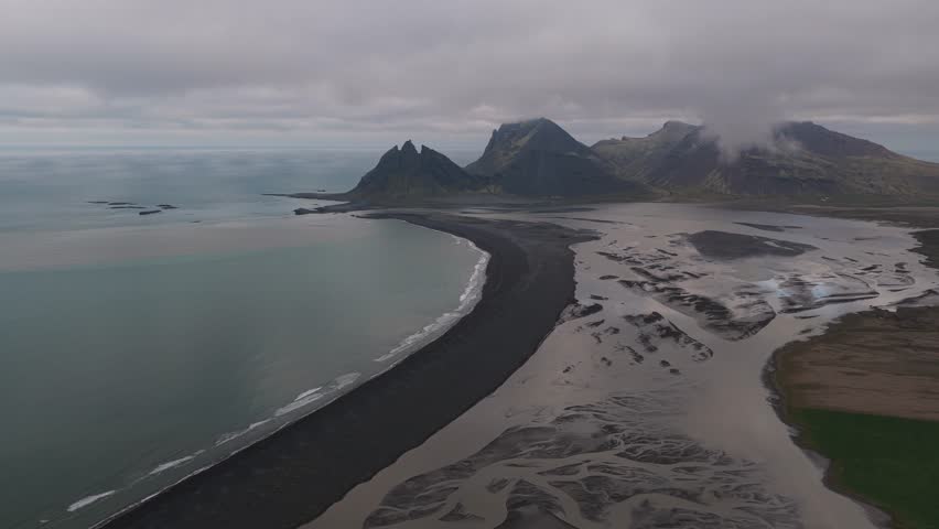 Aerial view of the stark black sand beach meeting the turquoise ocean near rugged mountains under a cloudy sky, Fjörur, Sveitarfélagið Hornafjörður, Iceland.