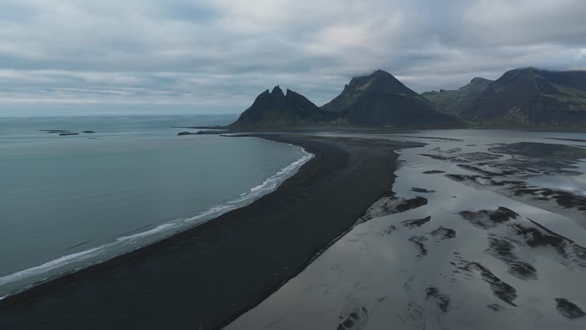 Aerial view of jagged mountain peaks meeting the dark volcanic sands and blue ocean under a cloudy sky, Fjörur, Sveitarfélagið Hornafjörður, Iceland.