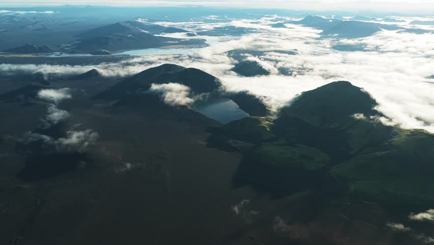 Aerial view of the rugged Hnausapollur Bláhylur View Point, where dark volcanic land meets green hills and a serene lake, Rangárþing ytra, Iceland.