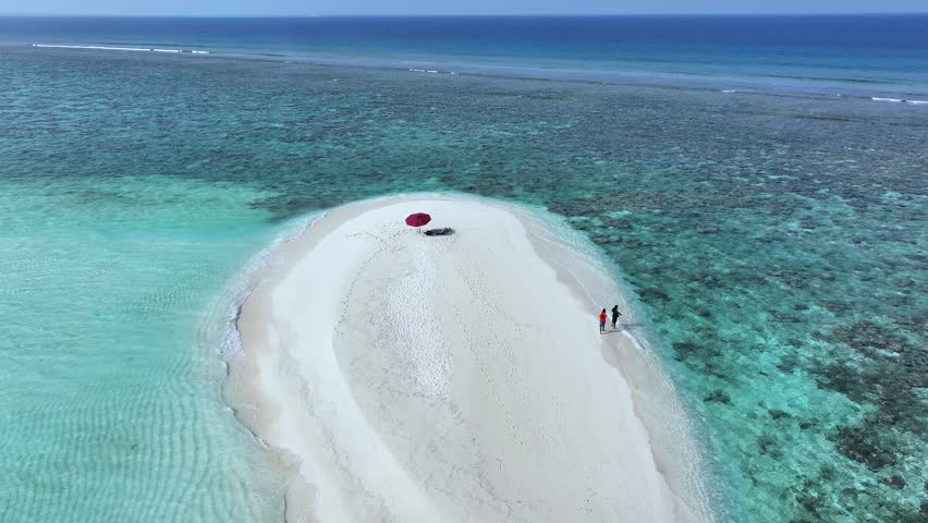 Aerial view of a pristine white sandbank meeting turquoise waters, with a red umbrella and couple enjoying the scenery, Thoddoo, Alif Alif Atoll, Maldives.