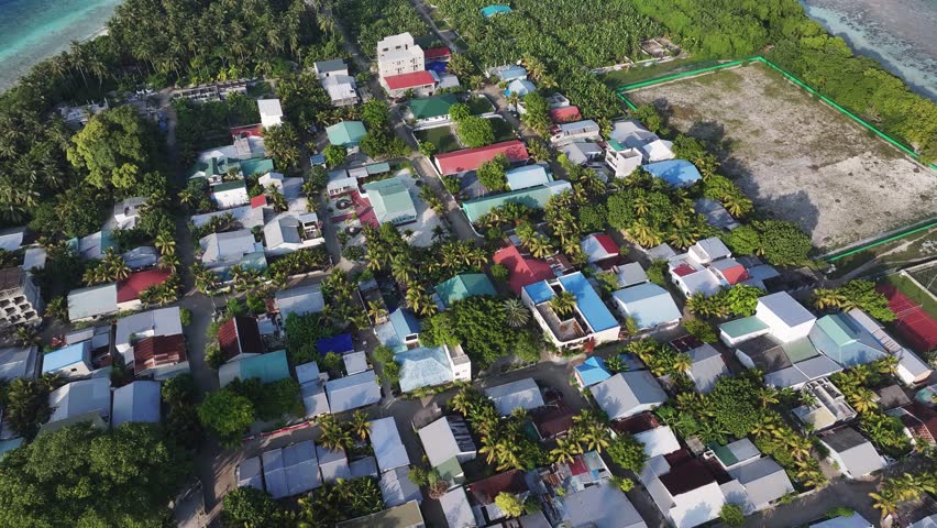 Aerial view of the island, a tapestry of white buildings nestled among lush green trees, fringed by turquoise waters and the expansive blue ocean, Kamadhoo, Baa Atoll, Maldives.