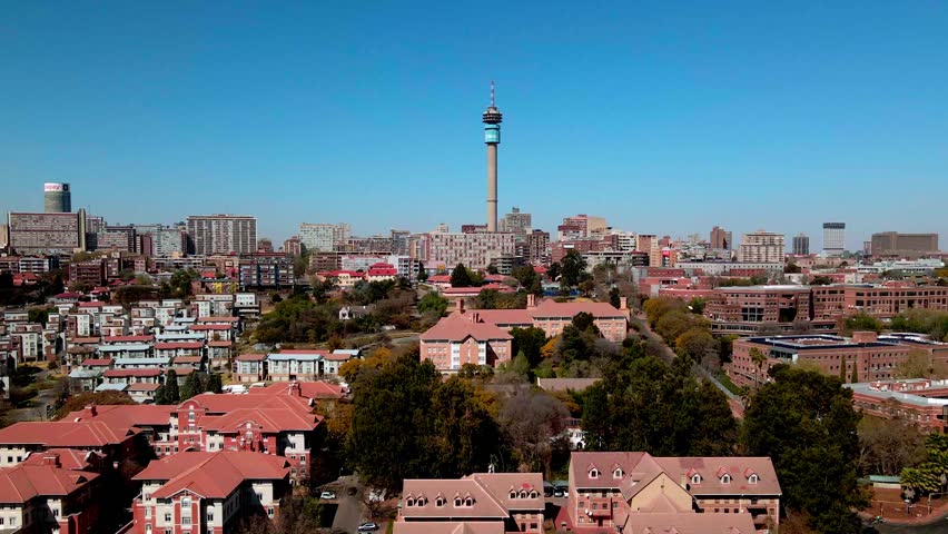 Aerial view of Hillbrow Tower, surrounded by buildings, trees, and streets, under a clear blue sky, Johannesburg, Gauteng, South Africa.
