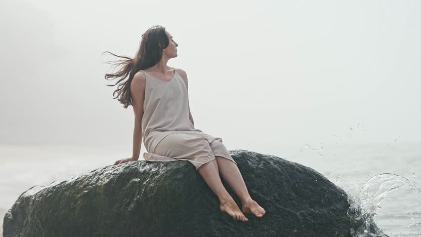 Young woman with dark hair sits barefoot on large rock at baltic sea coast, surrounded by dense fog. Soft light and muted tones create serene, contemplative atmosphere