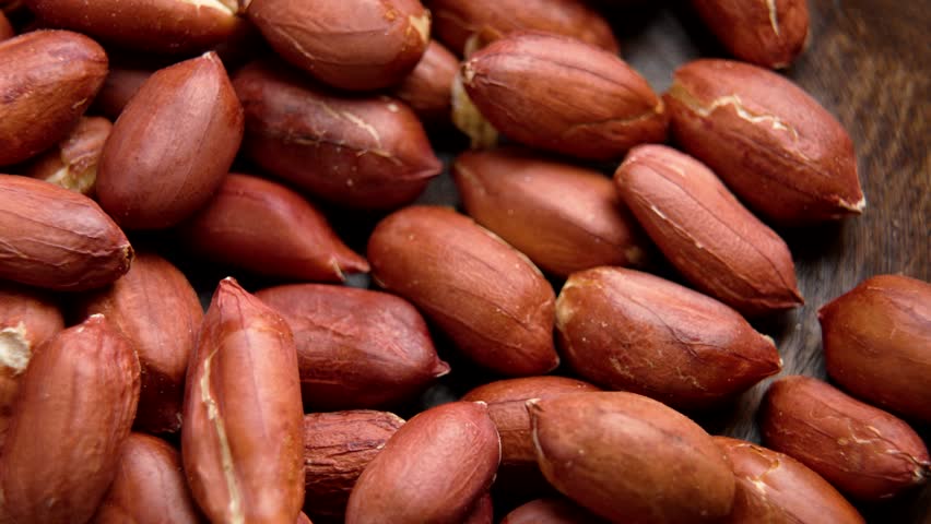 Metal spoon scoops shelled brown-skinned peanuts from wooden bowl. Top view. Macro