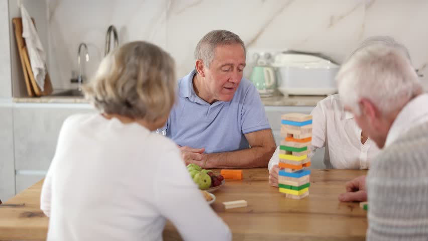 Elderly happy people playing Jenga at home, woman is removing a wooden block from the tower and the others are watching