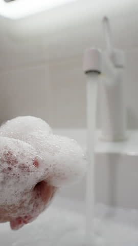 Close-up sequence showing hands being washed at a bathroom sink, from foamy soap lather to a thorough rinse under running water, emphasizing proper hygiene and cleanliness.