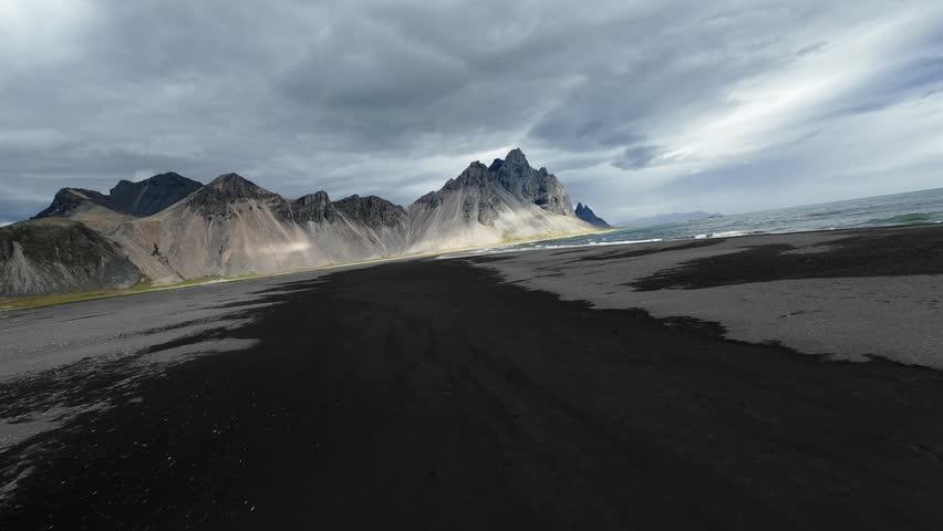 A cinematic FPV drone flight over the perfect black sand beach of Stokksnes, Iceland, flying directly towards the dramatic peaks of Vestrahorn mountain.