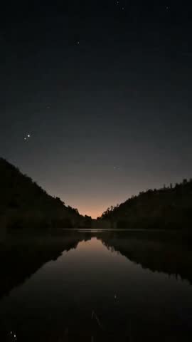 Serene twilight over a calm lake reflecting the dark silhouettes of hills under a faint starry sky.