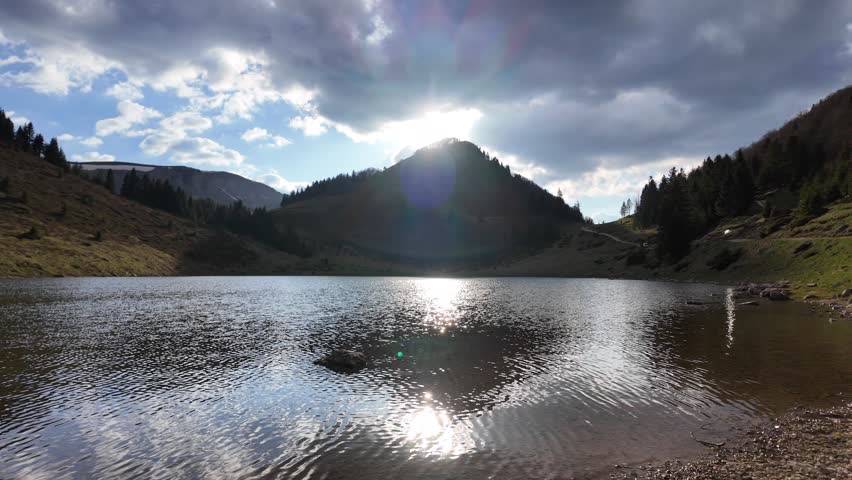A mountain lake landscape illuminated by bright sunlight with vivid water reflections at sunset, as the sun dips behind the mountains.